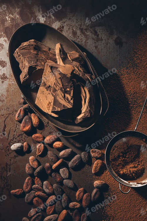 Preview: top view of delicious chocolate pieces, cocoa beans, powder and sieve on dark surface