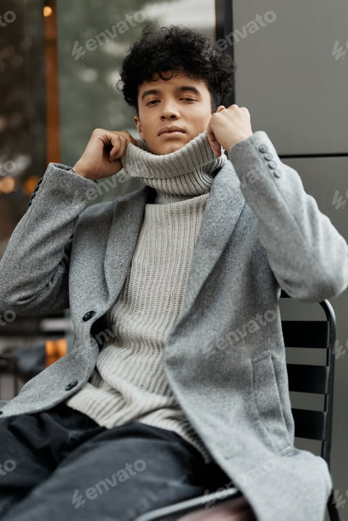 Preview: Fashionable young man in a grey sweater and jacket sitting on a chair with hands on ears and head