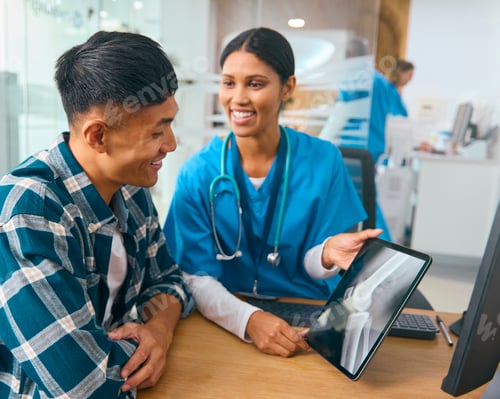 Preview: Female Doctor Wearing Scrubs At Hospital Appointment With Male Patient Looking At Digital Tablet