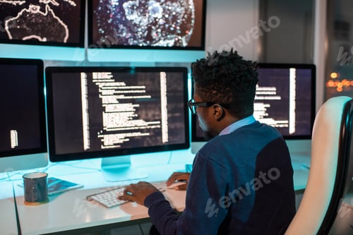 Preview: Young African software developer working late in front of computer screen with coded data