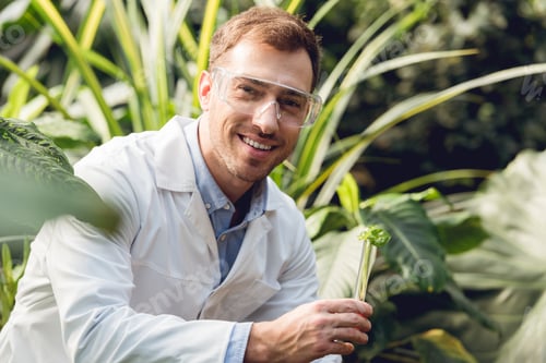Preview: selective focus of smiling handsome scientist in white coat and goggles taking plant sample in flask