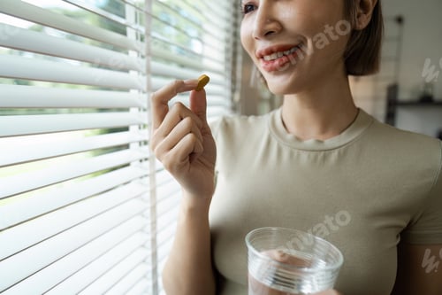 Preview: Smiling Woman Holding a Pill and Glass of Water by Window Blinds in Bright Room - Concept of