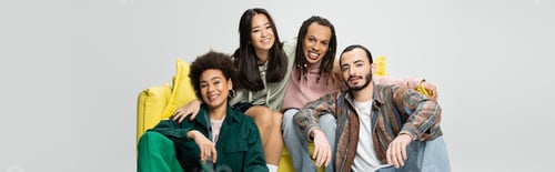 Preview: happy multicultural friends in trendy outfit looking at camera near yellow couch isolated on grey,