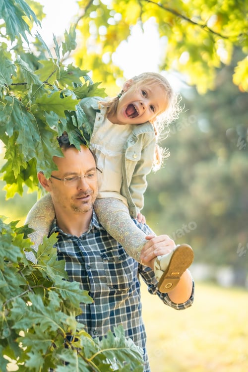 Preview: Young father and funny little daughter having fun together under tree in autumn park at sunset light