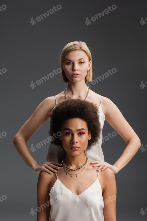 Preview: stylish interracial women in white strap dresses and colorful makeup looking at camera isolated on