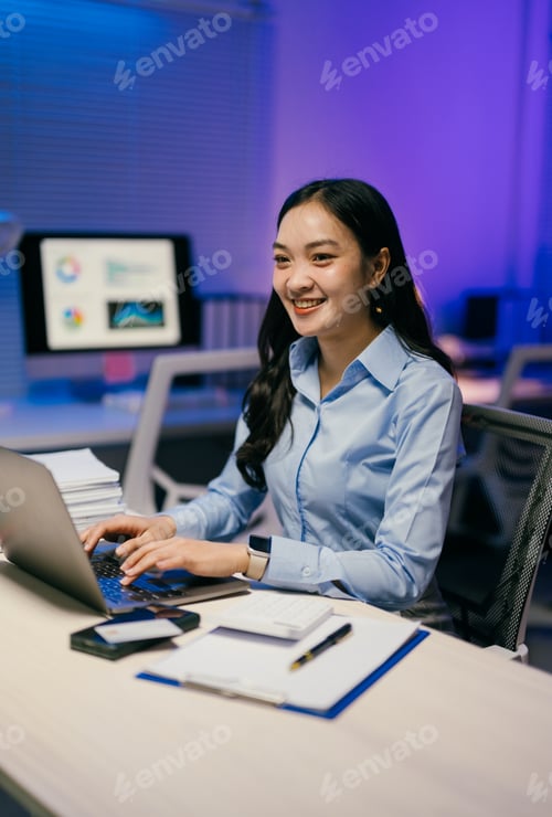 Preview: Young asian businesswoman smiling and working on a laptop at her desk in a modern office at night