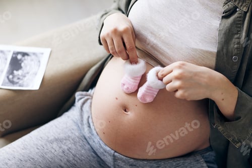 Preview: Hands of young pregnant female holding tiny socks for her future baby over belly