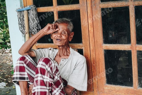 Preview: Thoughtful senior elderly man wearing saroong sitting in front of house with hands placed on head.