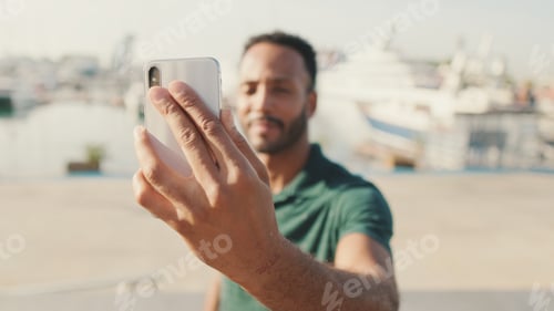 Preview: Young man takes selfie on the phone while sitting in harbor with yachts on the background