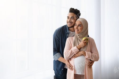 Preview: Portrait Of Happy Pregnant Islamic Couple Standing Near Window At Home