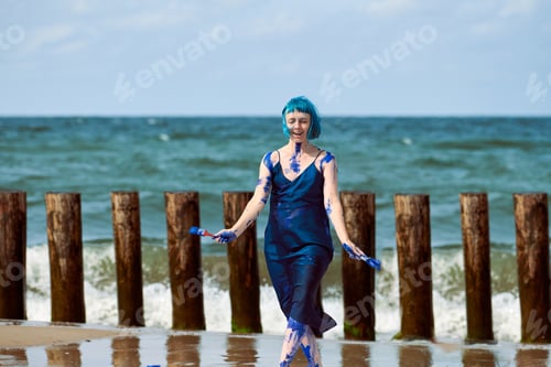Preview: Artistic blue-haired woman performance artist smeared with blue gouache paints dancing on beach