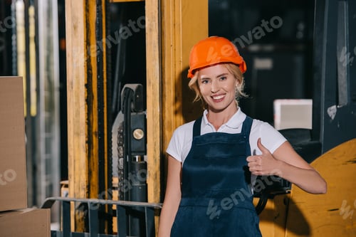 Preview: cheerful workwoman looking at camera and showing thumb up while standing near forklift loader