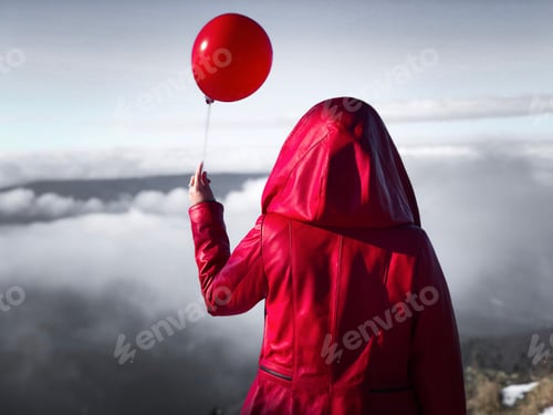 Preview: Woman in red hooded coat standing on top of a cliff with a red balloon and looking at the clouds