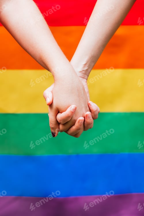 Preview: Close up of two woman's hands holding each other with the LGBTQ rainbow flag in the background