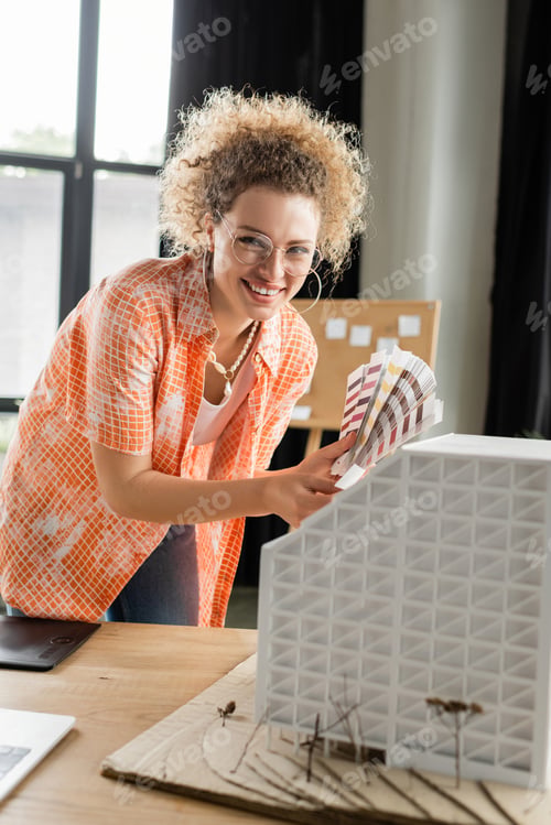 Preview: happy architectural designer in glasses holding color palette near residential house model in office