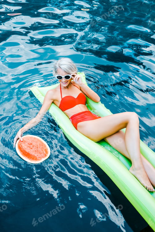 Preview: smiling blonde woman in swimsuit relaxing on green inflatable mattress in swimming pool with
