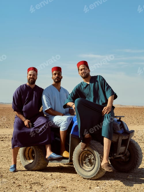 Preview: Three men wearing traditional fez hats pose confidently on a blue quad bike in a desert setting.