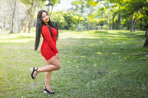 Preview: young latina female student, posing sideways in a red dress in a green park surrounded by nature.