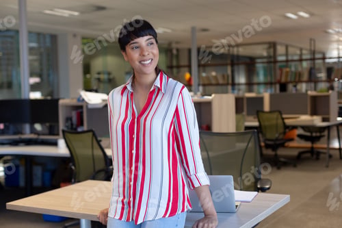 Preview: Thoughtful smiling biracial female advisor looking away while leaning on desk at modern workplace