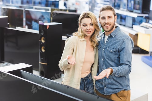 Preview: boyfriend and smiling girlfriend pointing with hands at new tv in home appliance store