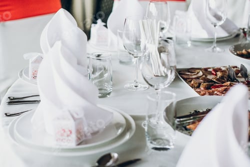 Preview: Wedding table with food, silver cutlery, empty glasses, white silk chairs and napkins