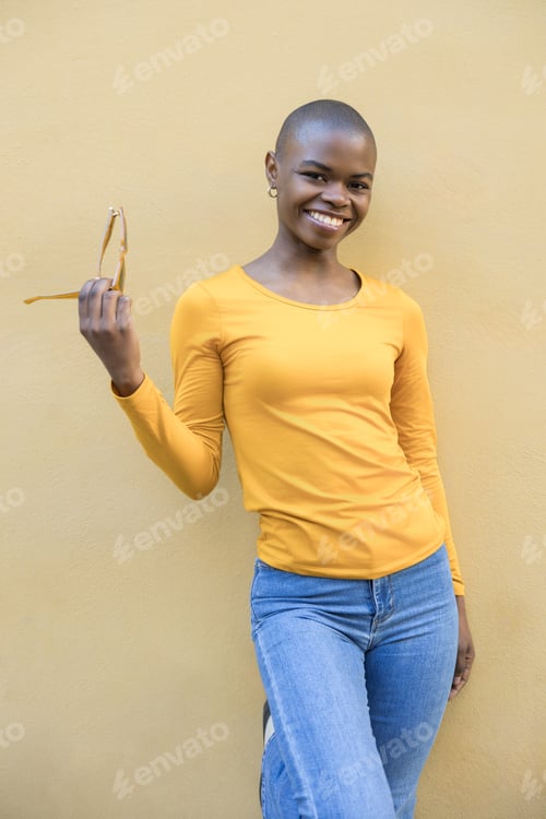 Preview: Smiling Black woman with a shaved head, dressed in yellow and blue jeans, posing stylishly