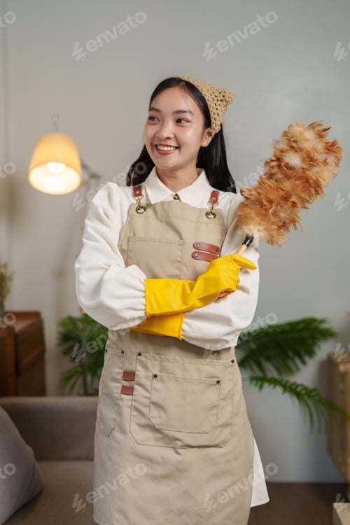 Preview: Smiling young housekeeper wearing yellow gloves and an apron holds a feather duster while standing i