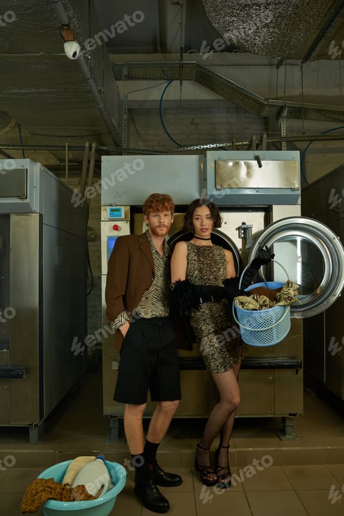 Preview: interracial couple in stylish outfits posing near clothes and washing machines in public laundry