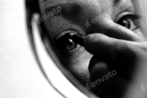 Preview: Grayscale shot of a female applying cosmetics to an upper eyelid in front of the mirror