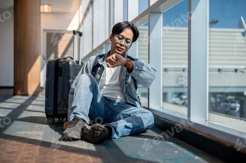 Preview: Asian man sitting on floor looking at time on watch waiting to depart or missed a flight in airport