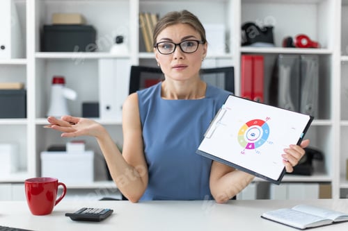 Preview: A young girl sits at a table in the office and holds a pen and a document with a diagram.