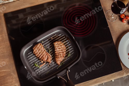 Preview: Top view of grilled steaks with rosemary in frying pan on induction stove