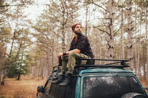 Preview: Millennial man hipster with beard sitting on car roof drinking beer in a pine forest.
