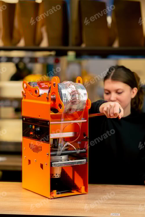 Preview: close-up an orange machine for packaging drinks with plastic on a bar counter girl using the machine