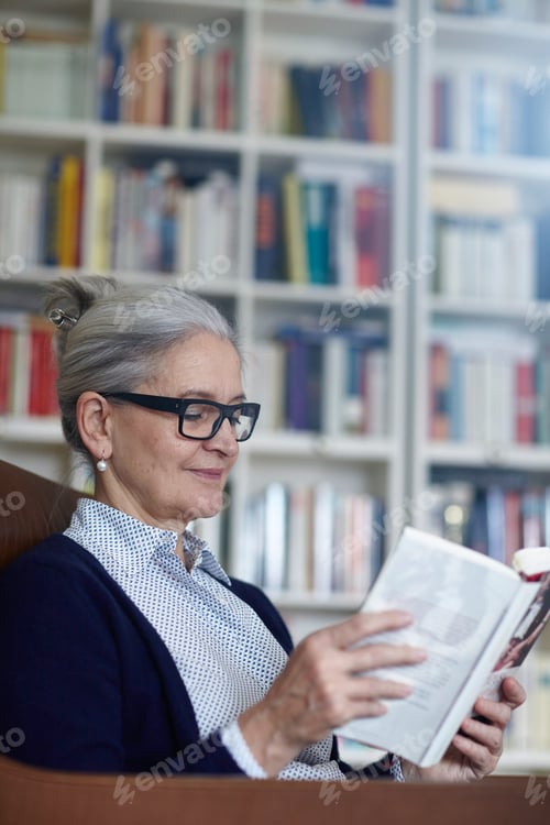 Preview: Grey haired mature woman reading book from bookshelves