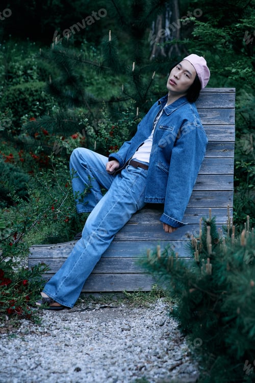 Preview: Stylish young man relaxing in a park wearing denim outfit and cap on a sunny day