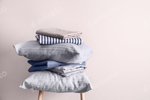 Preview: Stack of clean bed sheets and pillows on stool near beige wall. Space for text