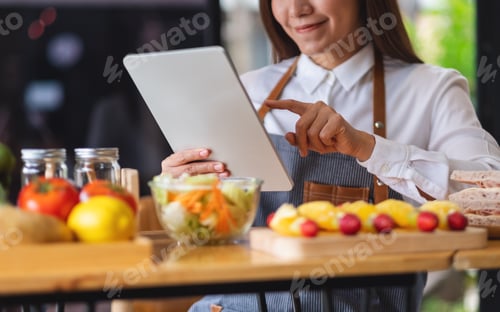 Preview: A beautiful young asian woman following recipe on digital tablet while cooking salad and sandwich