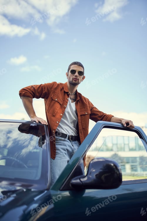 Preview: vertical shot of appealing young man in stylish vibrant clothing posing next to his car, fashion