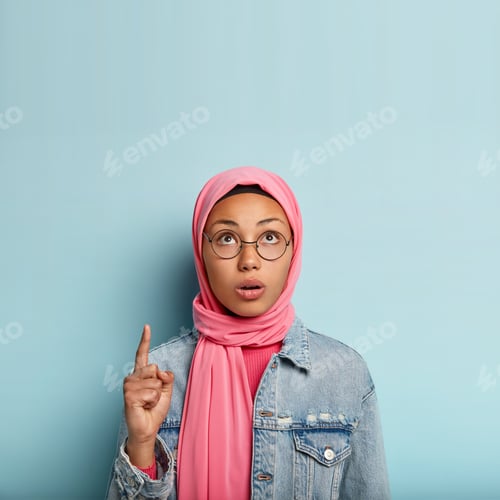 Preview: Surprised young Muslim woman points up with one fore finger, focused upwards, has concerned facial e