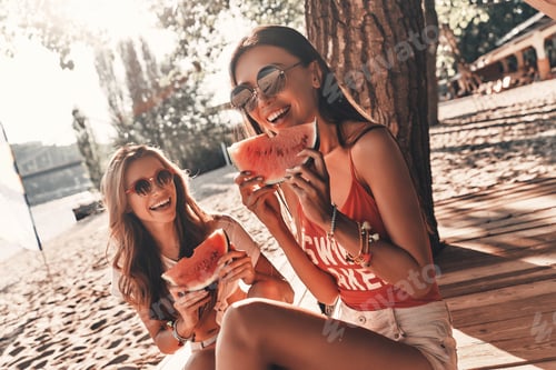 Preview: Refreshing. Two attractive young women smiling and eating watermelon while sitting on the beach