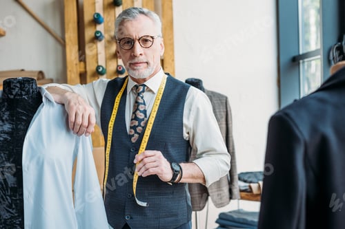 Preview: handsome tailor leaning on mannequin at sewing workshop and looking at camera