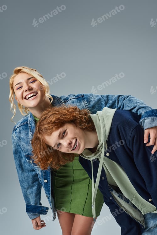Preview: laughing blonde teen girl embracing stylish redhead boyfriend while having fun in studio on grey