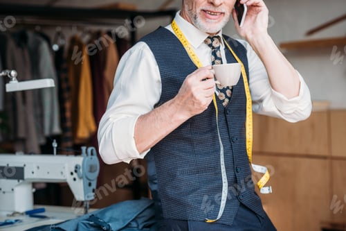 Preview: cropped image of tailor standing with cup of coffee and talking by smartphone at sewing workshop