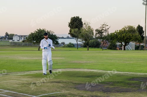 Preview: Male cricketer wearing white kit and pads standing on worn pitch at dusk holding wooden bat