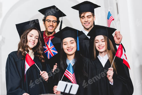 Preview: selective focus of cheerful students in graduation gowns holding flags of different countries and