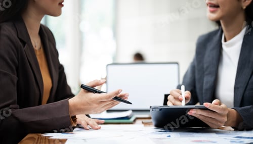 Preview: Two business women sit at desk discuss project details, working on graph chart and laptop computer