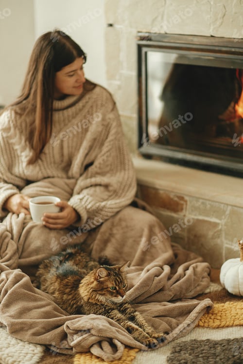 Preview: Cute cat and woman with cup of tea sitting together on cozy blanket at fireplace