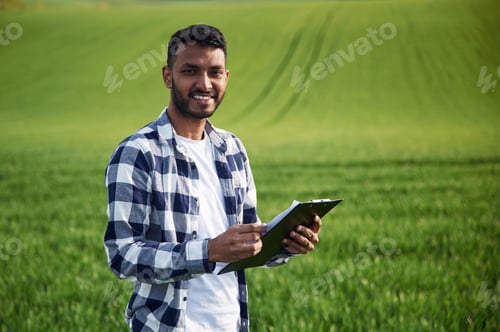 Preview: Standing and holding notepad. Handsome Indian man is on the agricultural field