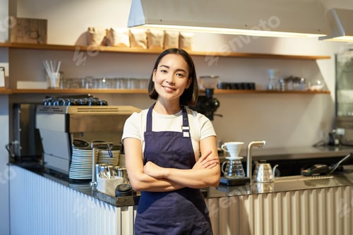 Preview: Smiling asian girl barista, standing near counter in cafe, wearing company apron, cross hands and